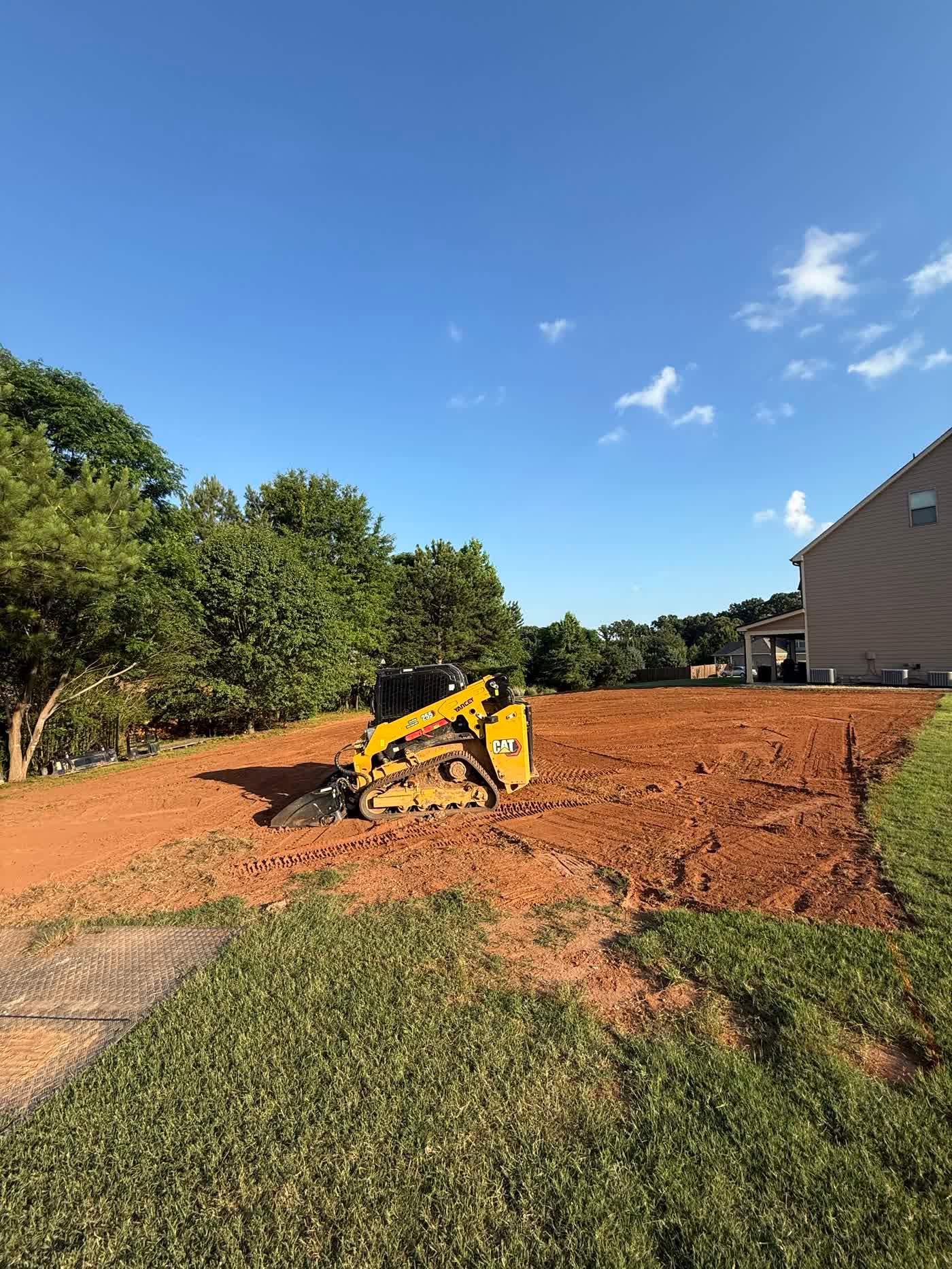 Skid steer cutting grade across a red clay lot under blue sky